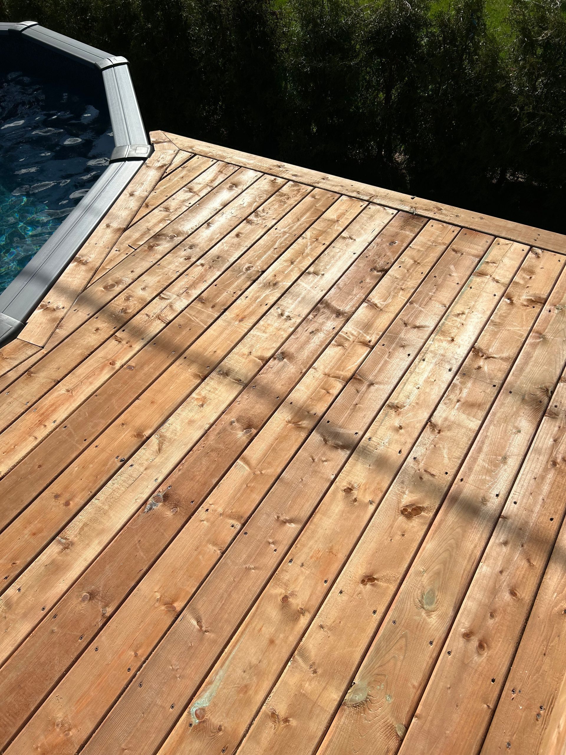 Terrasse en bois entourant une piscine à l'eau bleue, planches inclinées et végétation verte.