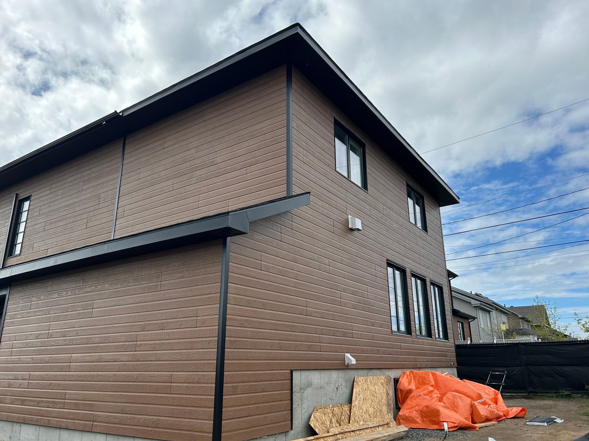 Maison brune à deux étages avec des boiseries noires sous un ciel nuageux. Matériaux de construction visibles.