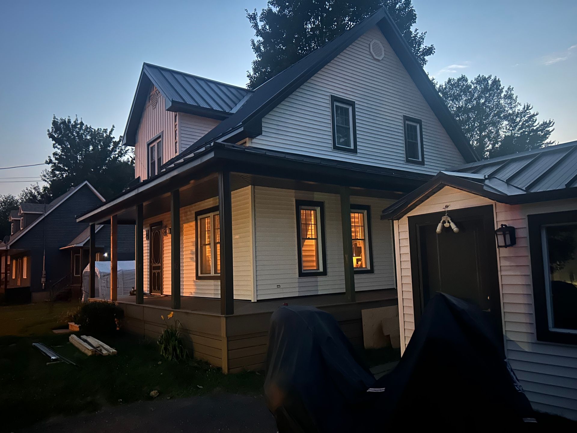 Maison blanche à deux étages avec un porche sombre, un toit en métal et des fenêtres éclairées au crépuscule.