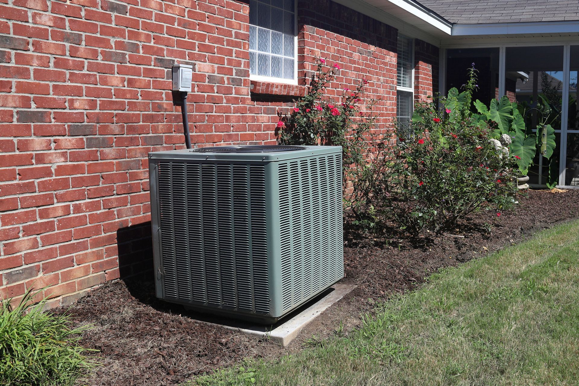 Outdoor HVAC unit beside a brick building, next to shrubs and grass.