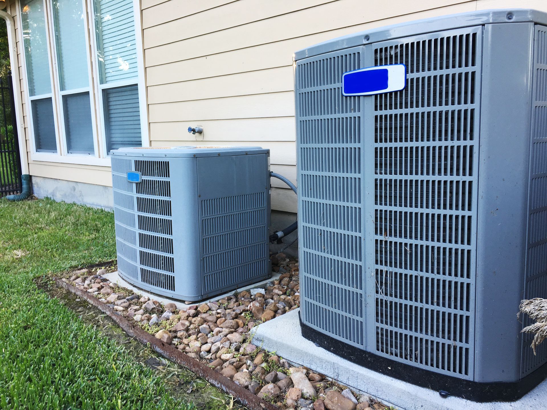 Two gray outdoor HVAC units beside a house wall and window, on gravel and concrete pads.