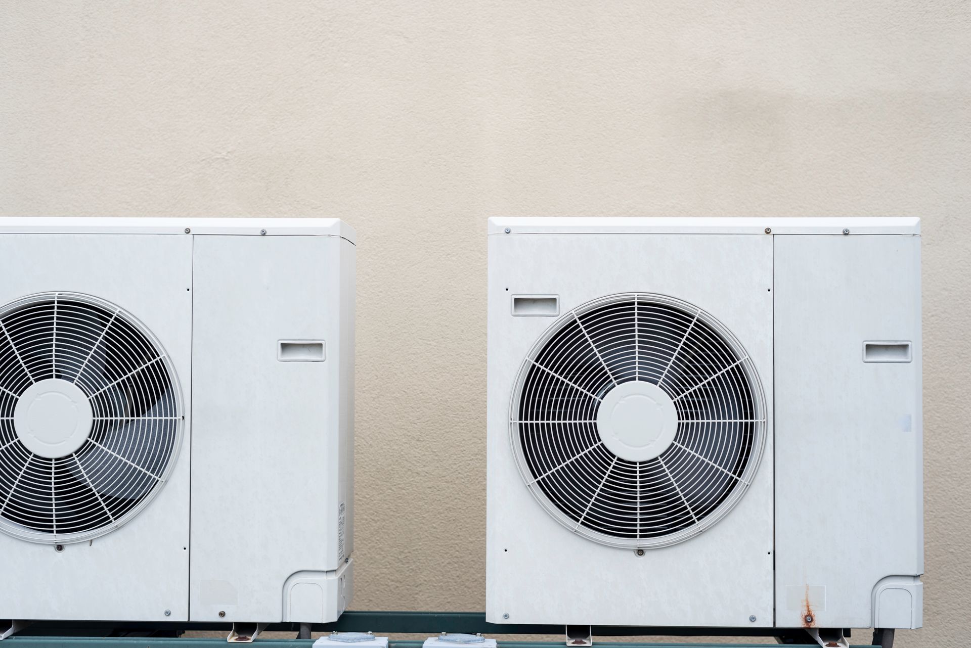 Two white outdoor air conditioning units against a beige wall