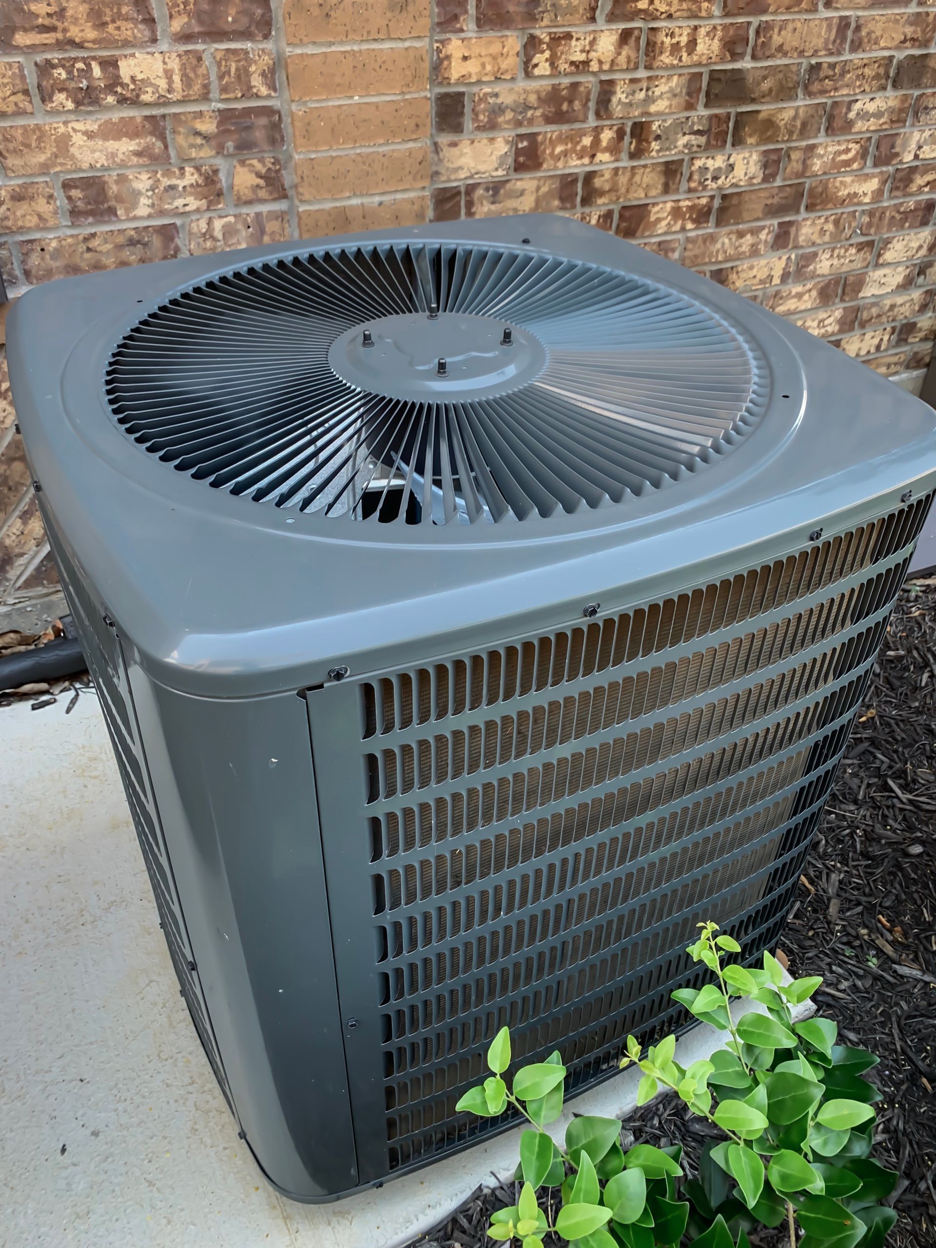 Outdoor air conditioner condenser beside a brick wall, with a fan grille and a small plant in front