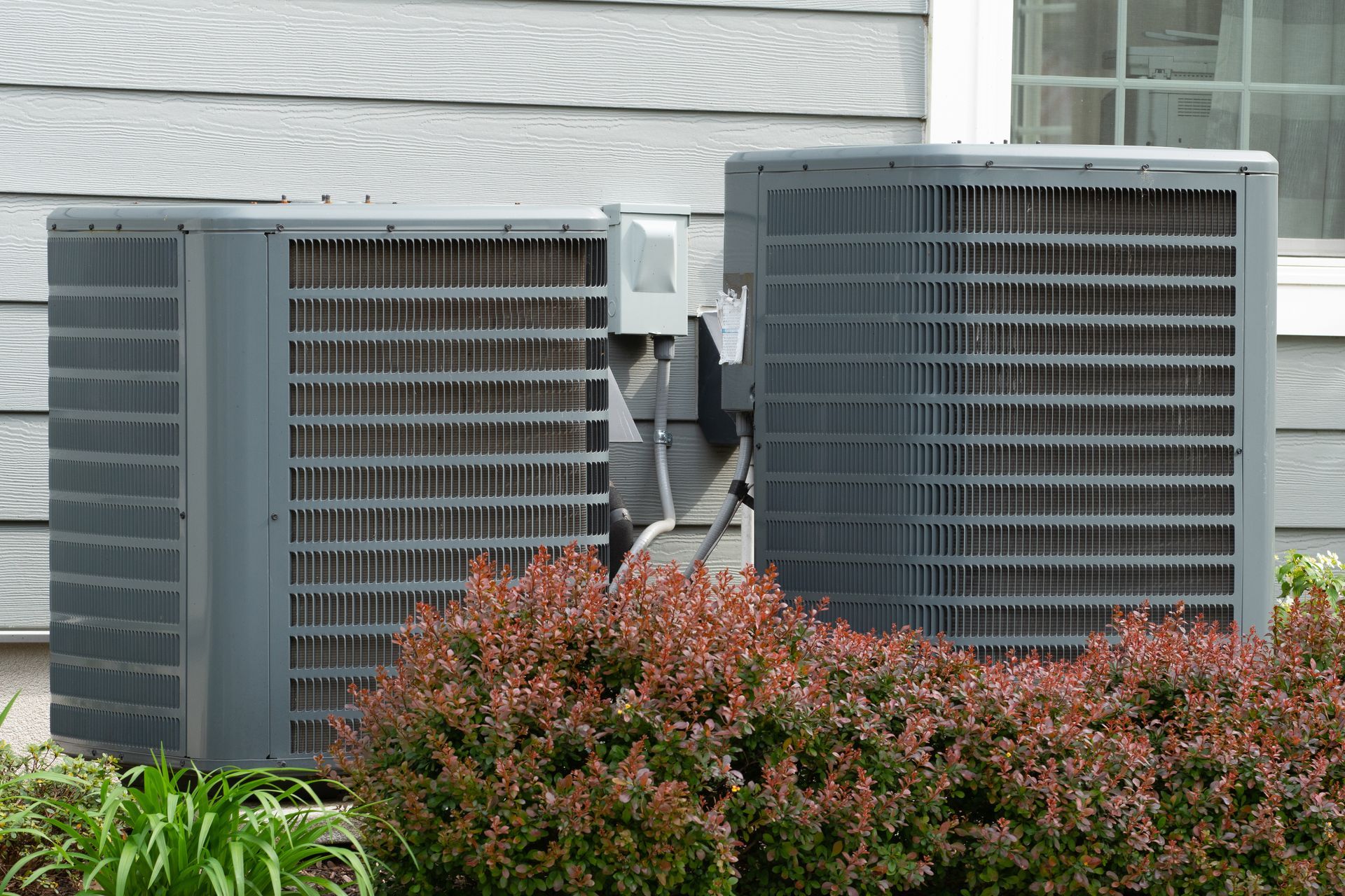 Two gray outdoor HVAC condenser units beside a house, partly hidden by shrubs.