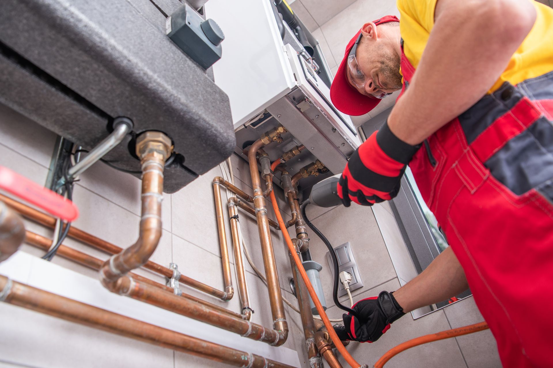 Technician in red overalls inspecting copper pipes and wiring under a boiler.