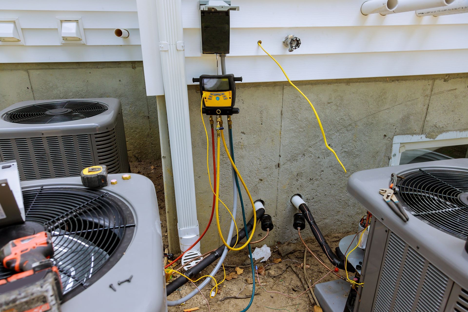Outdoor HVAC units with a yellow maintenance monitor and hoses on a concrete wall