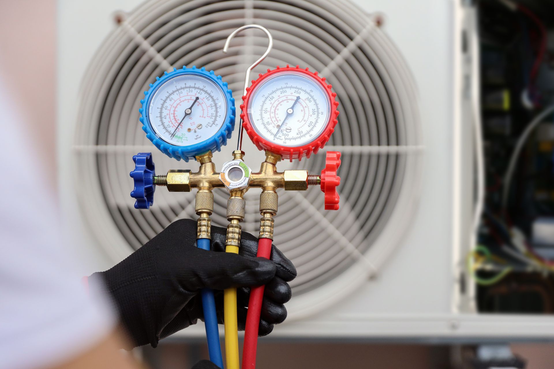 Gloved hand holding HVAC manifold gauges with blue and red dials in front of a fan unit