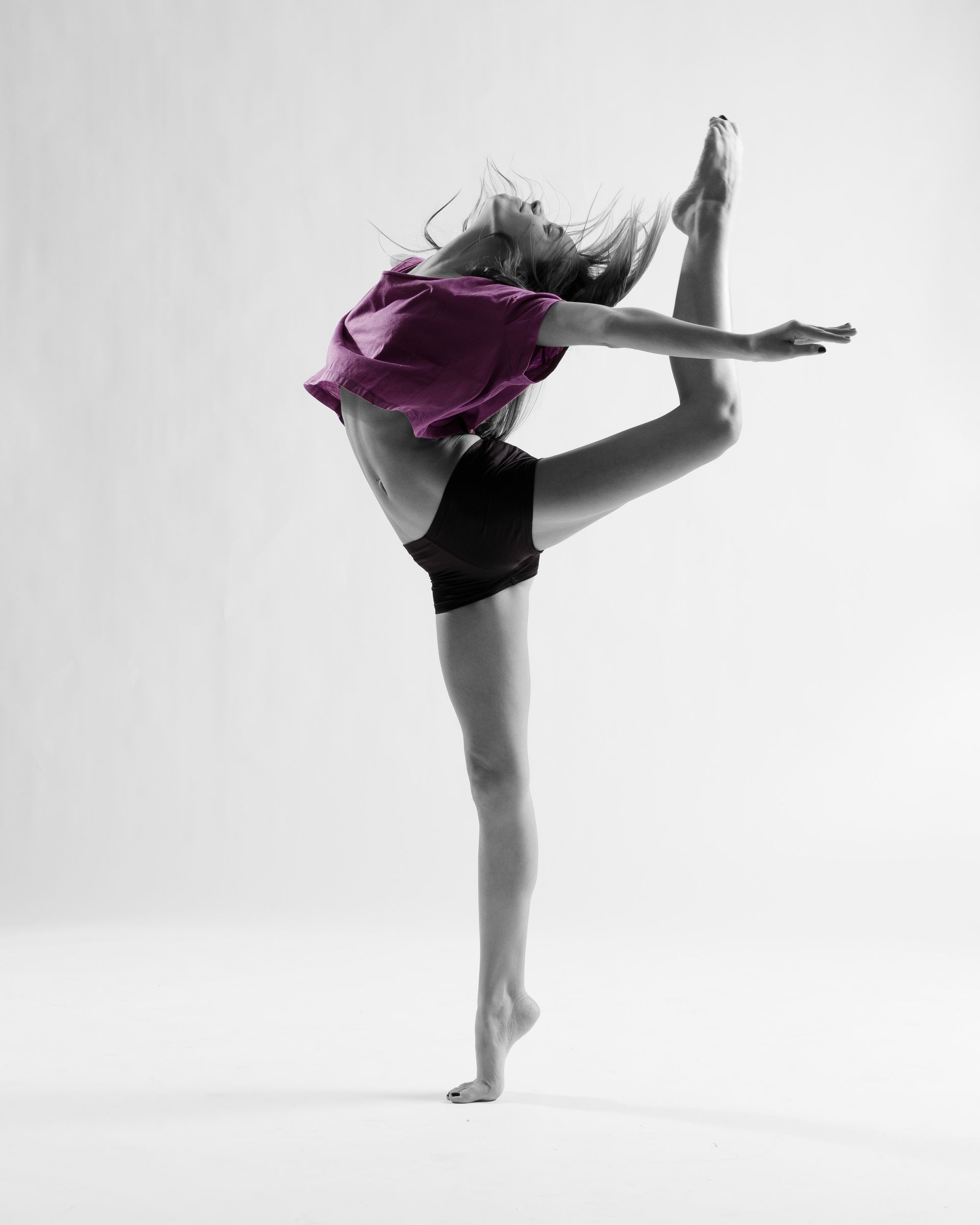 Dancer in mid-air, bending backward, holding one foot. Wearing purple shirt, black shorts, studio background.