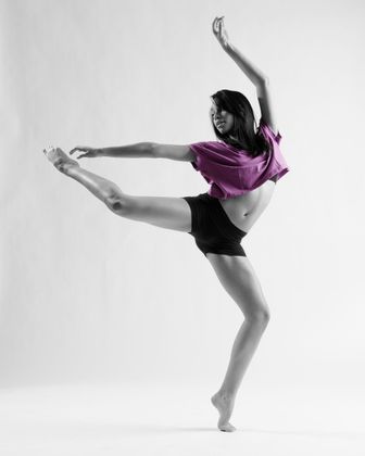 Dancer in a graceful pose, leg extended, arm raised, wearing a purple top and black shorts.