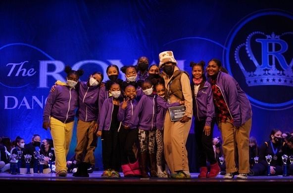 Dance group in purple hoodies on stage with trophy. Blue backdrop, logo with crown.