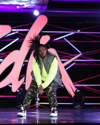 A dancer in neon green and camo pants on stage, performing a hip-hop move.
