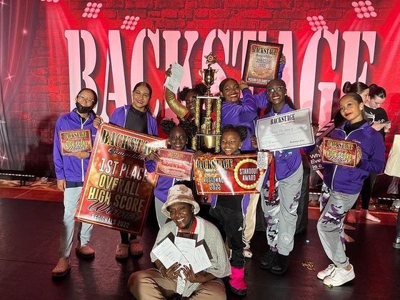 Dance team celebrates a win, holding trophies and awards on a stage. Purple outfits, happy expressions.
