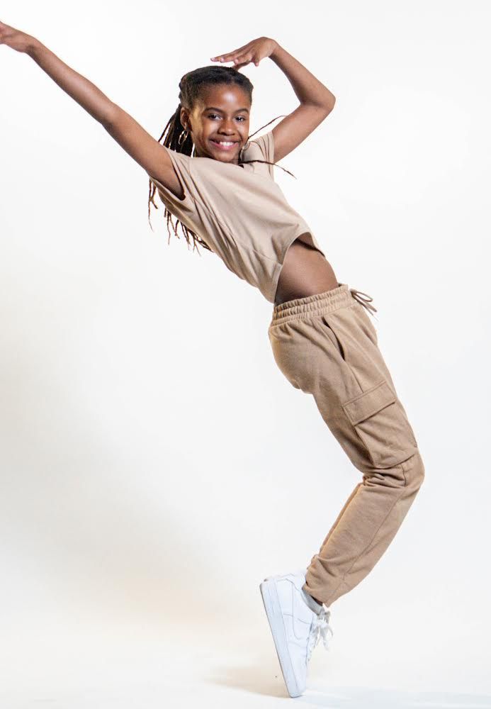 Young woman in tan outfit, dancing, smiling, reaching arms up and out, white sneakers, studio background.