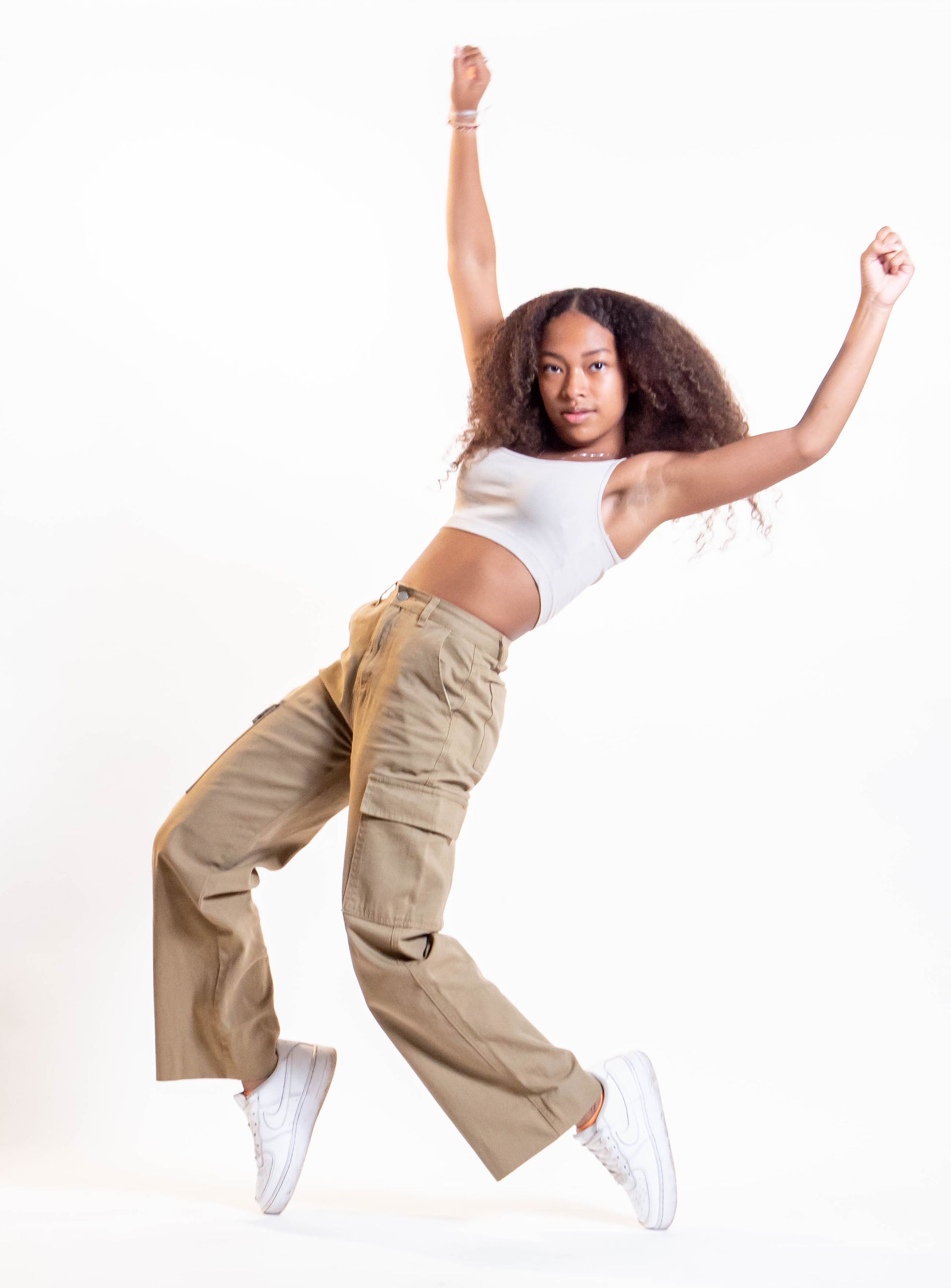 Young woman in dance pose, wearing white crop top, tan cargo pants, and white sneakers, arms raised, white background.