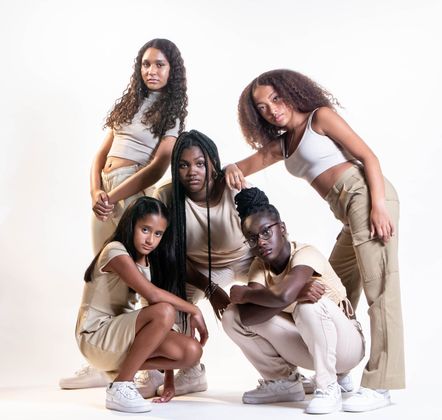 Five young women in beige outfits pose against a white backdrop; some crouch and smile.