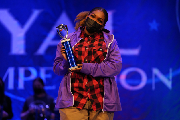 A masked woman in purple and red plaid, holding a trophy on stage at a competition.