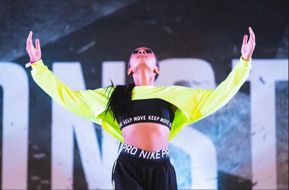 Dancer in neon yellow jacket, arms raised, athletic wear, against a stage backdrop.