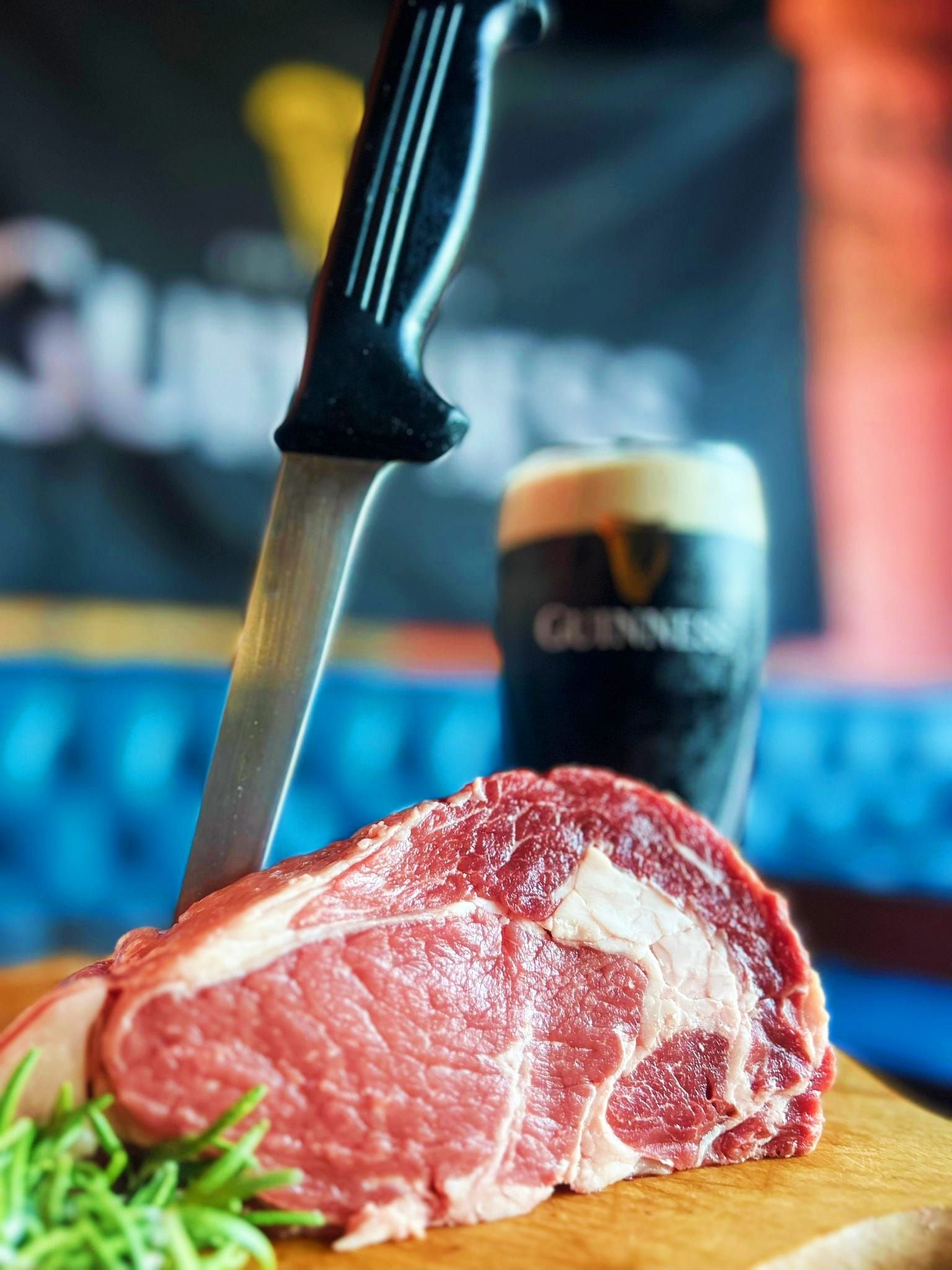 Bistecca cruda con un coltello infilato, rosmarino e una birra Guinness sullo sfondo.