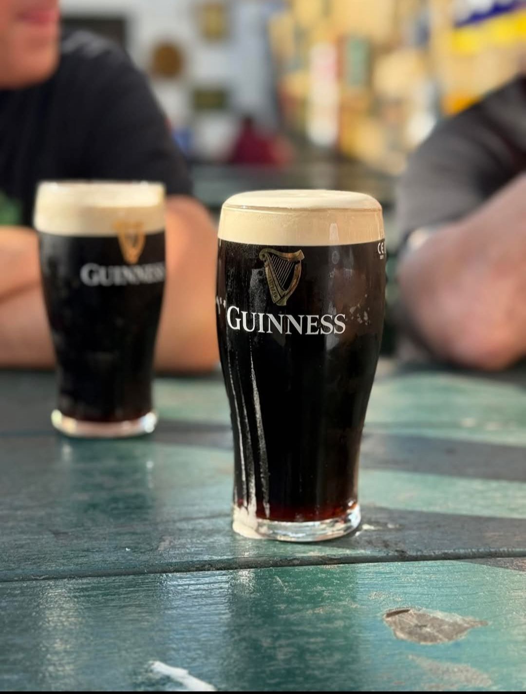 Due pinte di Guinness stout su un tavolo verde; una a fuoco, una birra scura con schiuma cremosa, l'altra sfocata.