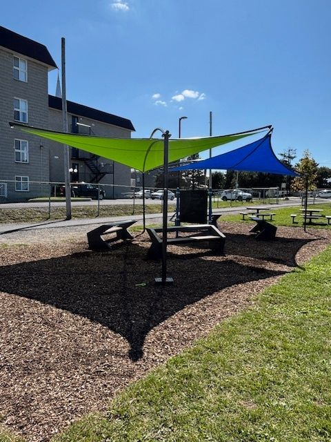Aire de pique-nique ombragée avec des auvents verts et bleus, des bancs et un bac à sable par une journée ensoleillée.