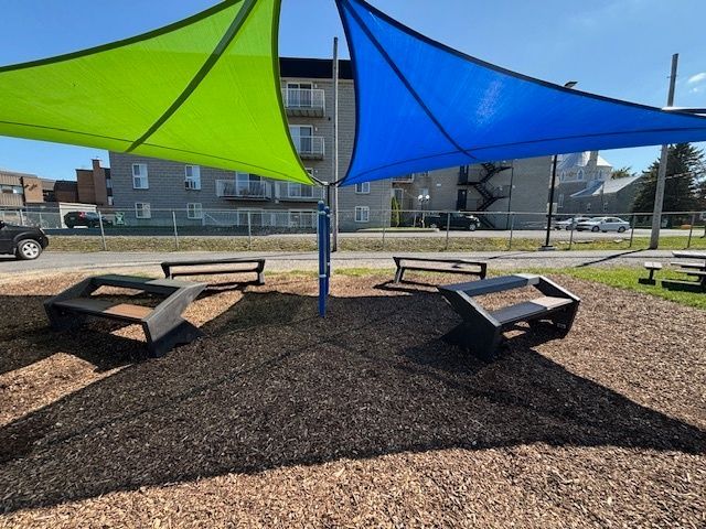 Des voiles d'ombrage vertes et bleues recouvrent des bancs noirs dans un parc recouvert de copeaux de bois.