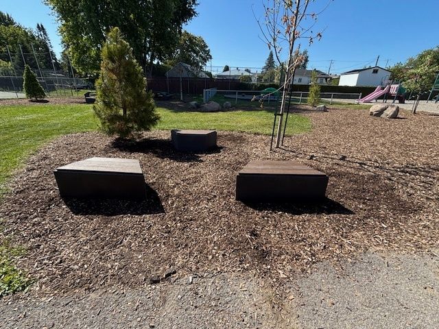 Trois bancs bruns entourés de copeaux de bois dans un parc. De l'herbe verte, des arbres et une aire de jeux se trouvent à l'arrière-plan.