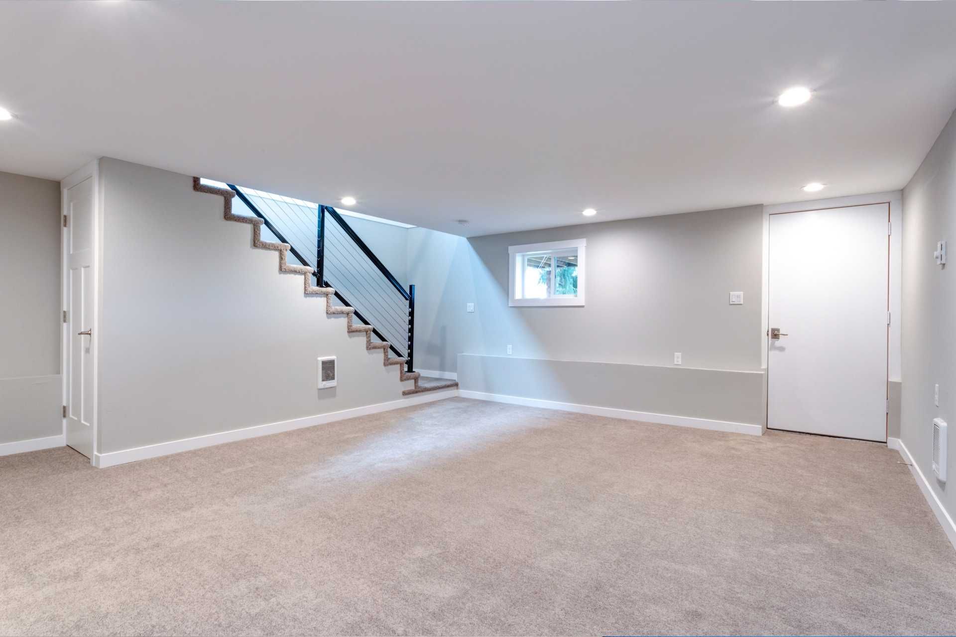 Empty carpeted basement room with stairs, small window, and white doors