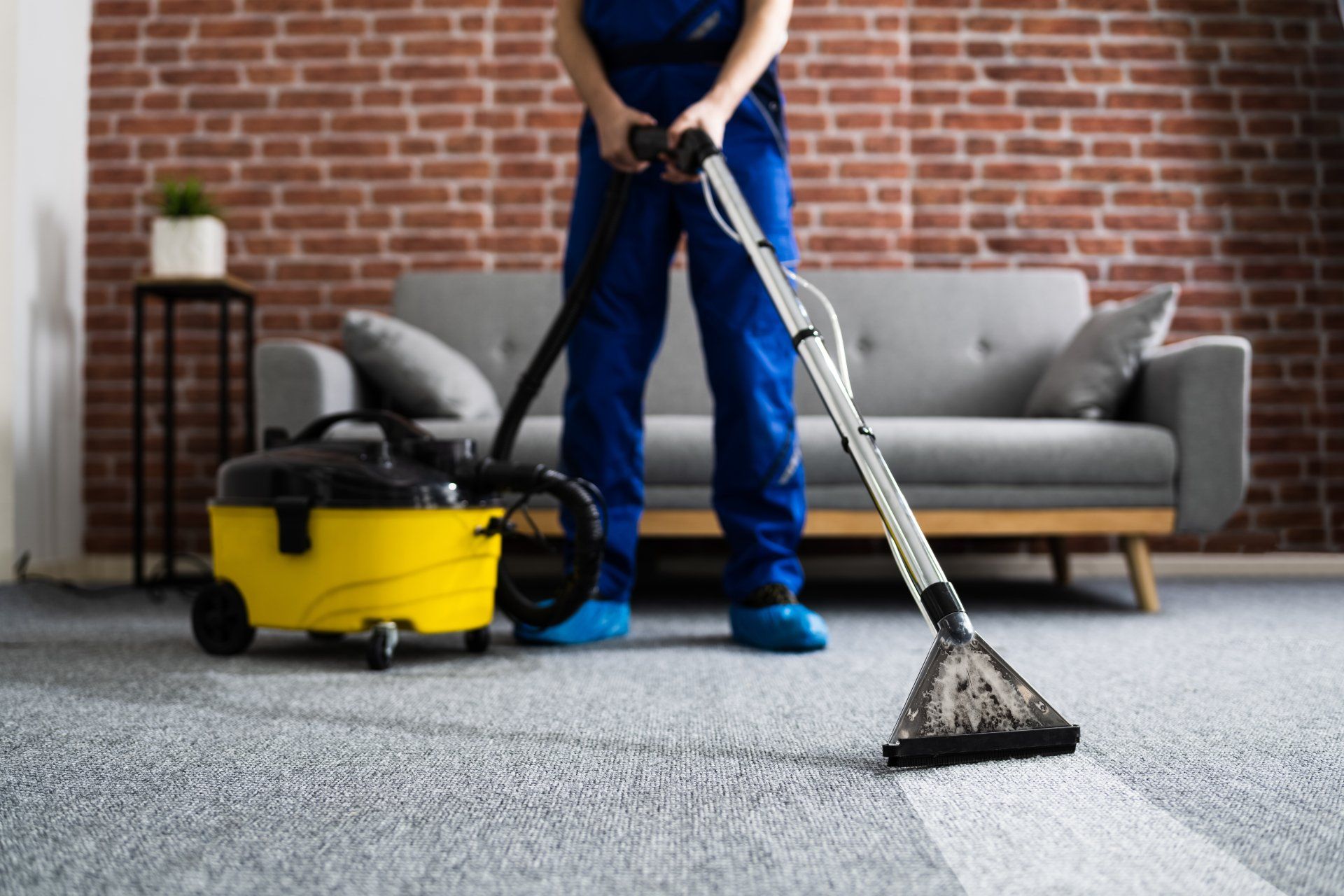 Person vacuuming a gray carpet in a living room with a yellow cleaner and brick wall background