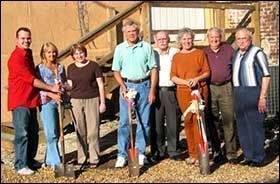 Eight people stand in a line outdoors holding shovels at a groundbreaking ceremony in front of a wooden building.