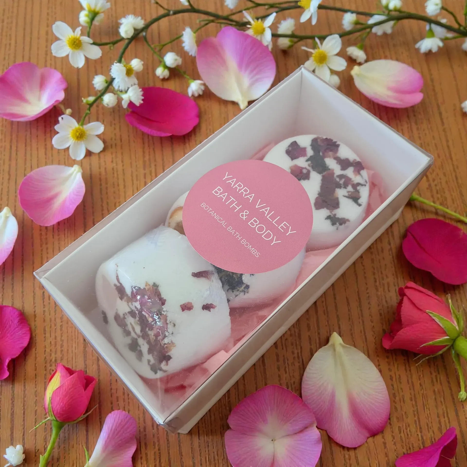 Pink and white floral bath bombs in a gift box on a wooden surface, surrounded by petals and daisies