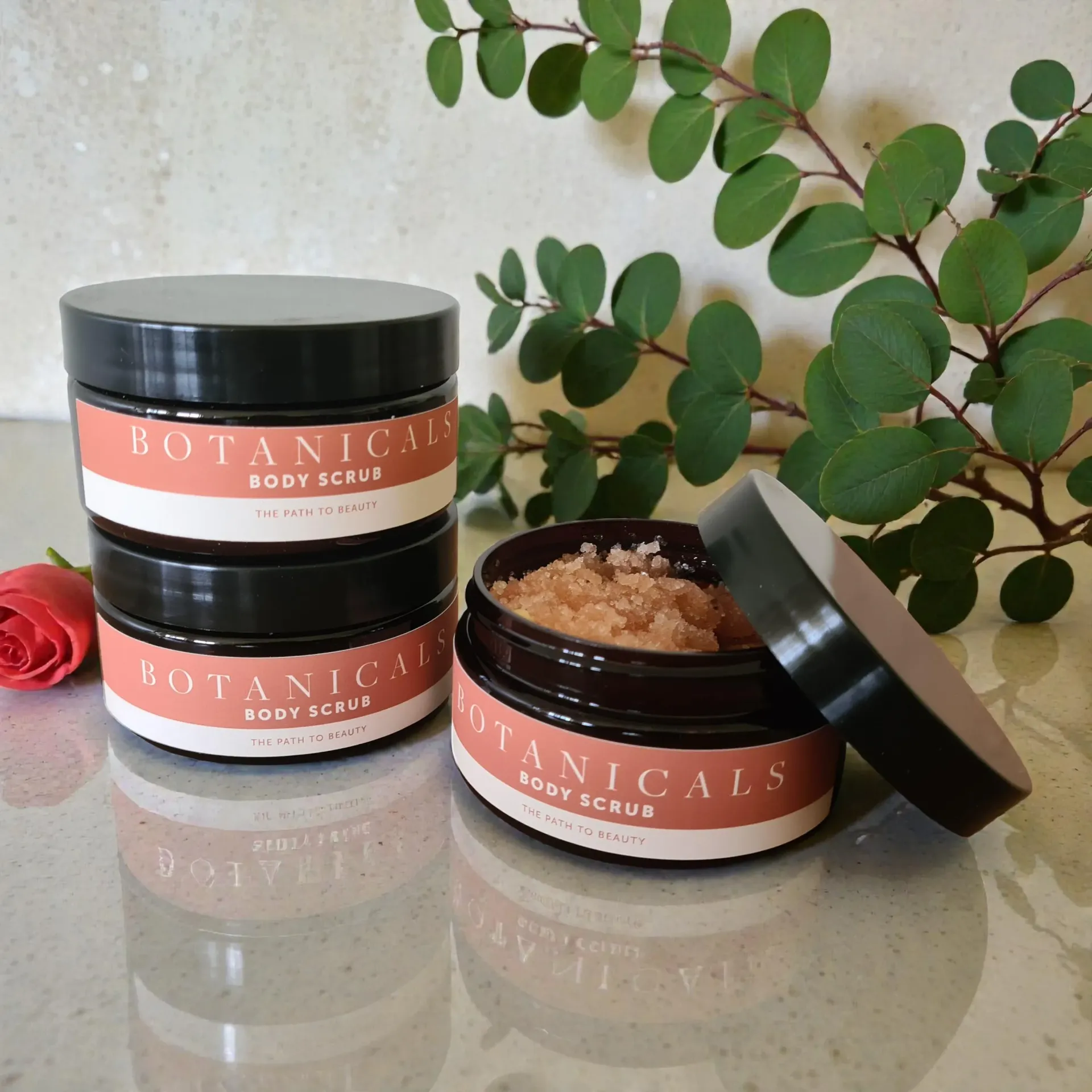 Botanicals body scrub jar with black lid among pink and green flowers
