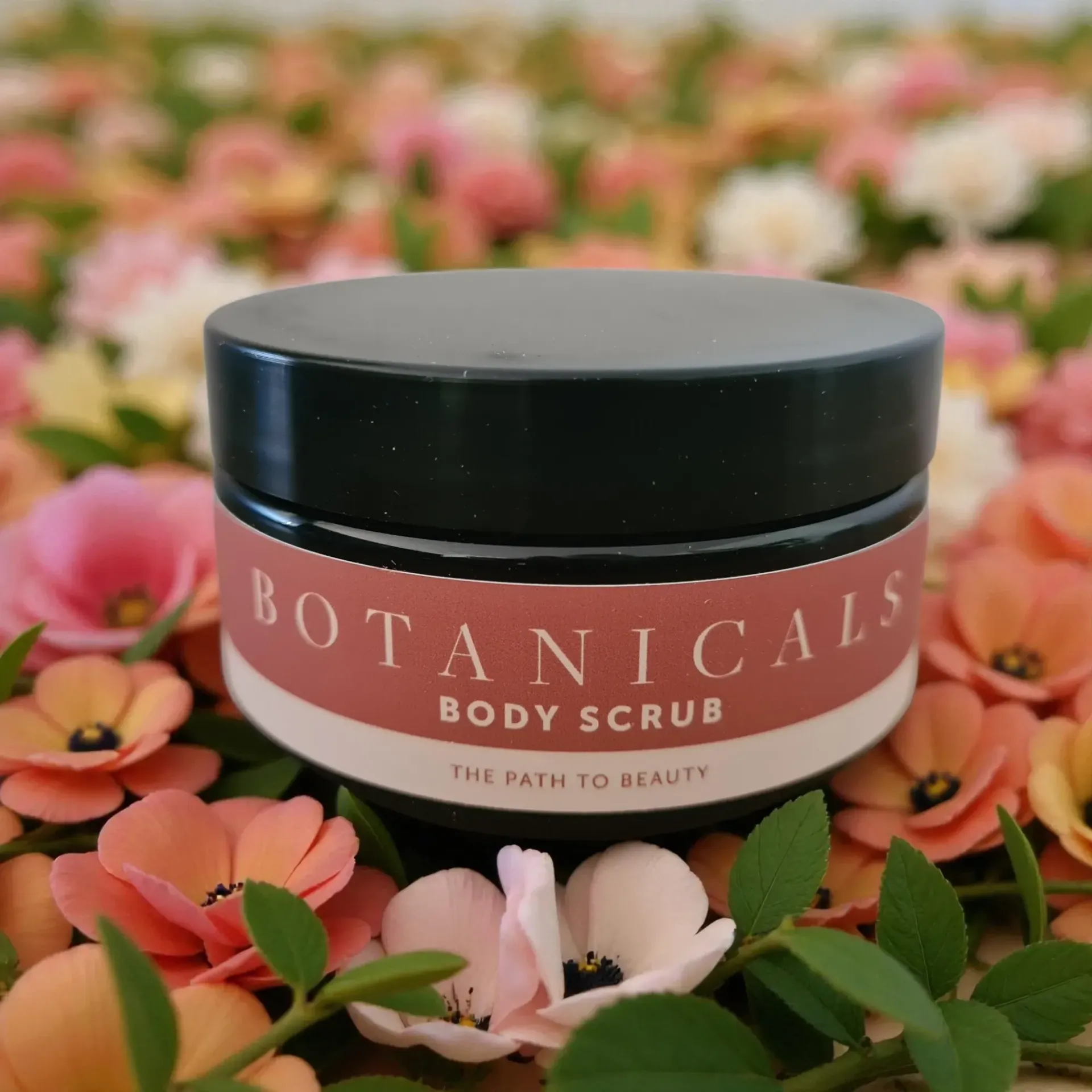 Botanicals body scrub jar with black lid among pink and green flowers