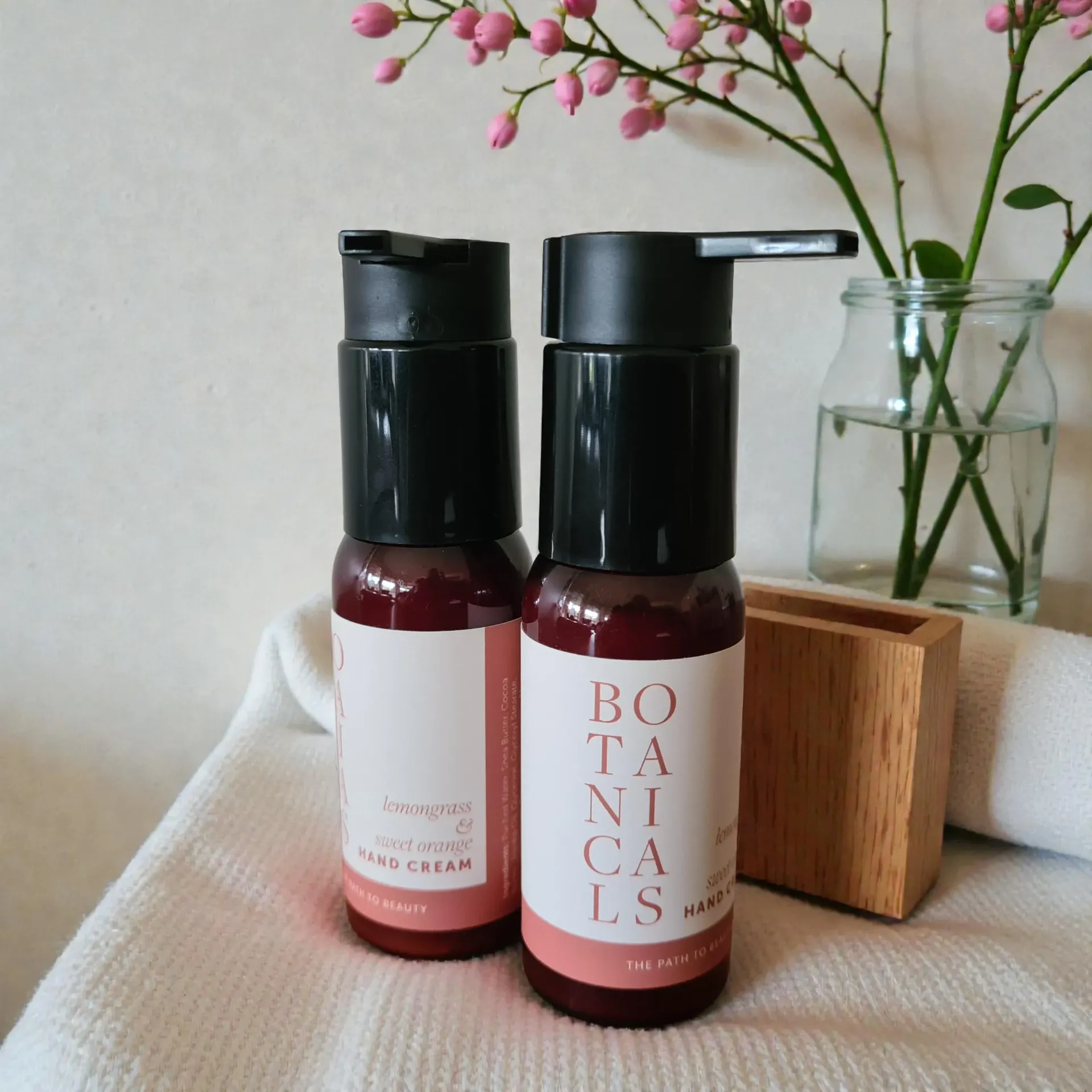 Two pink botanical skincare pump bottles of hand cream on a fabric surface beside flowers and a wooden block