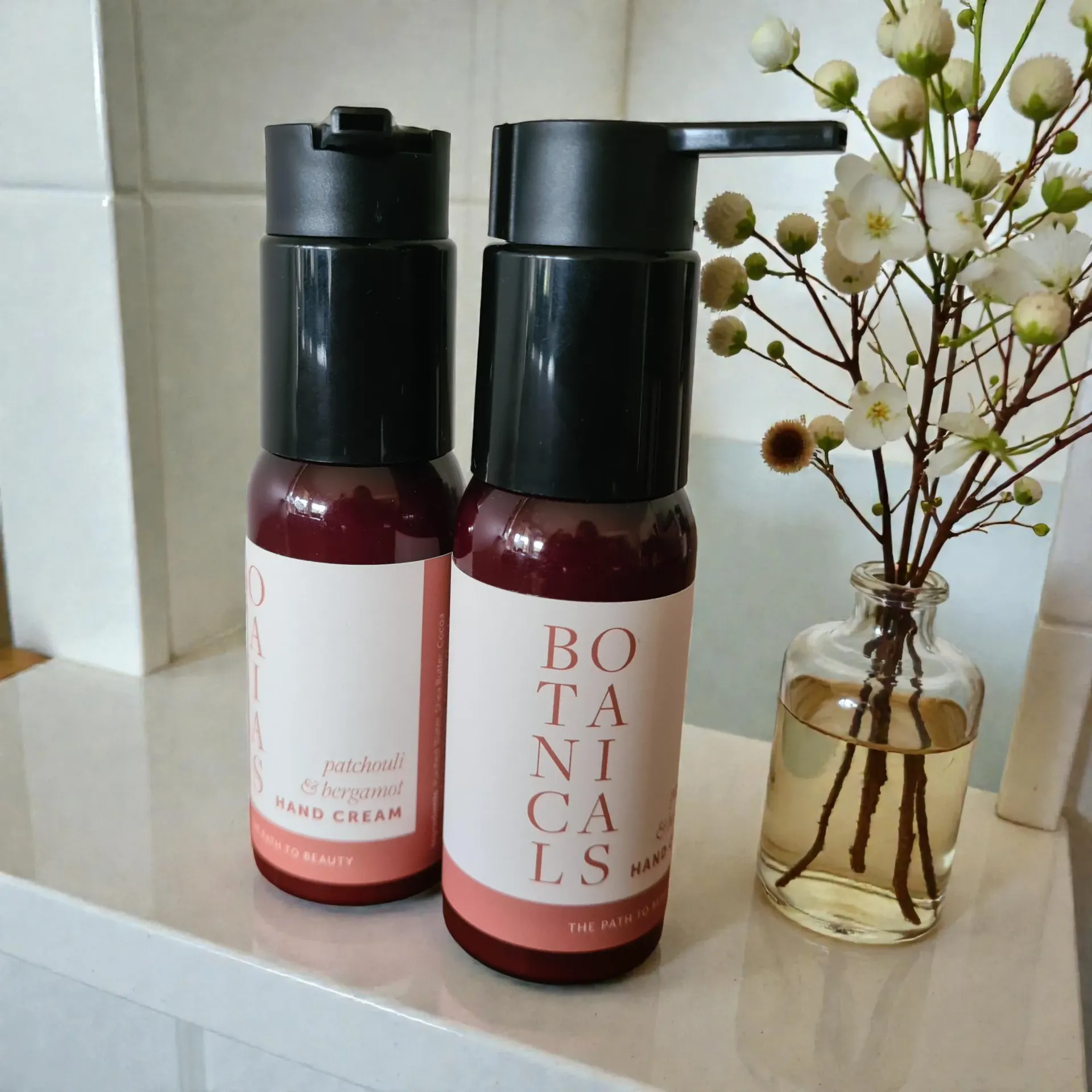 Two pump bottles of hand cream beside a small vase of dried flowers on a shelf