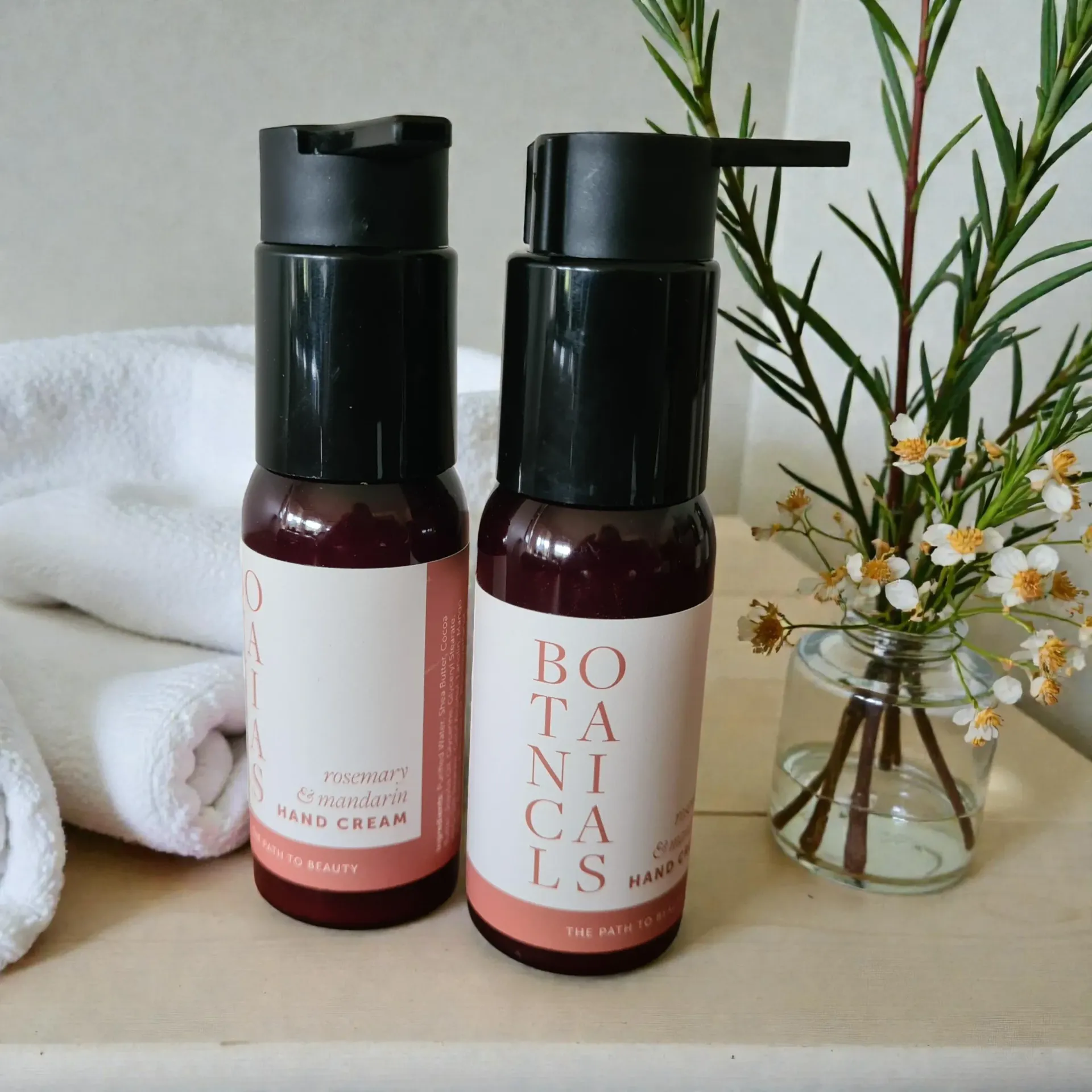 Two pink hand cream bottles on a towel beside a small vase of flowers and greenery