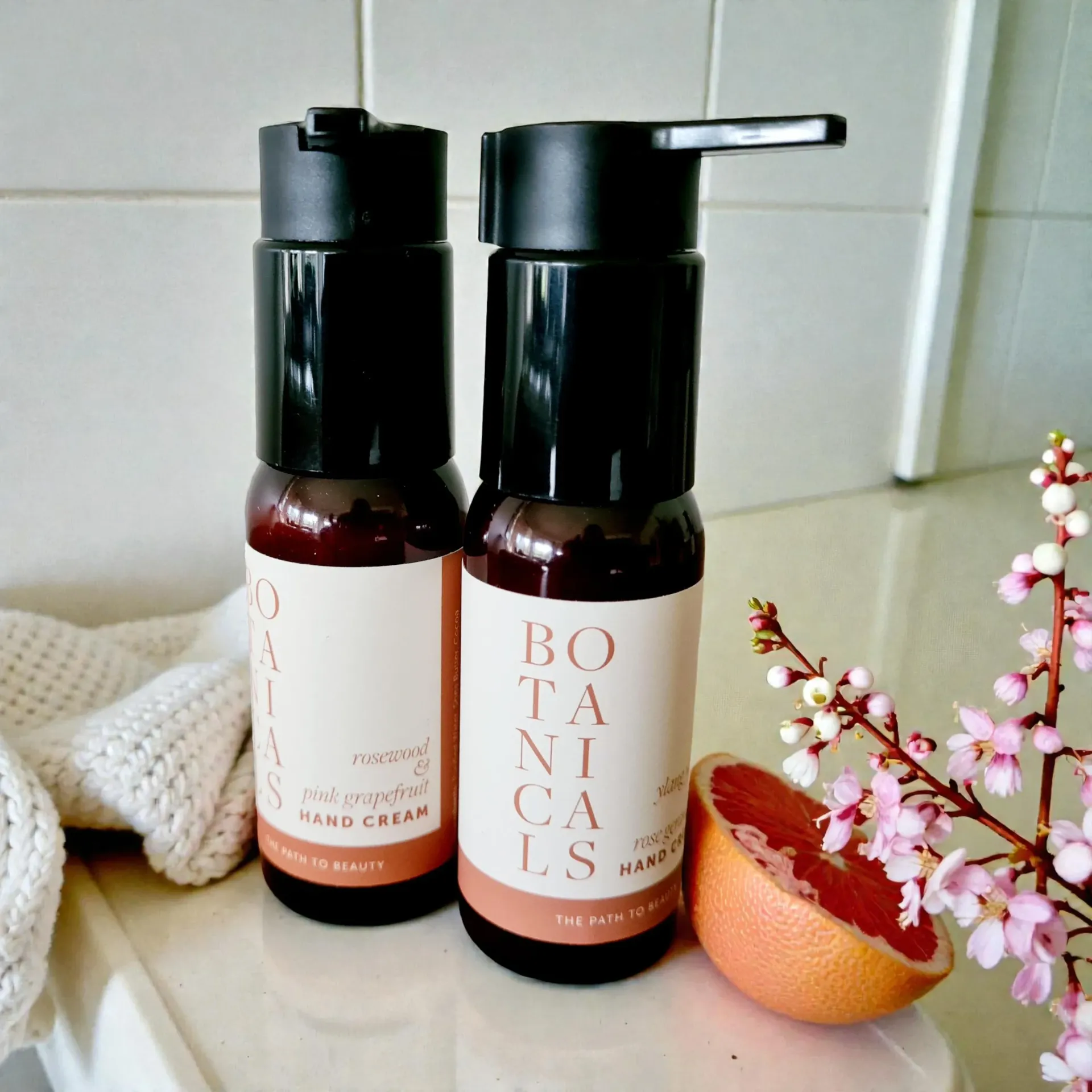 Two amber pump bottles of hand cream on a bathroom counter beside a sliced orange and flowers