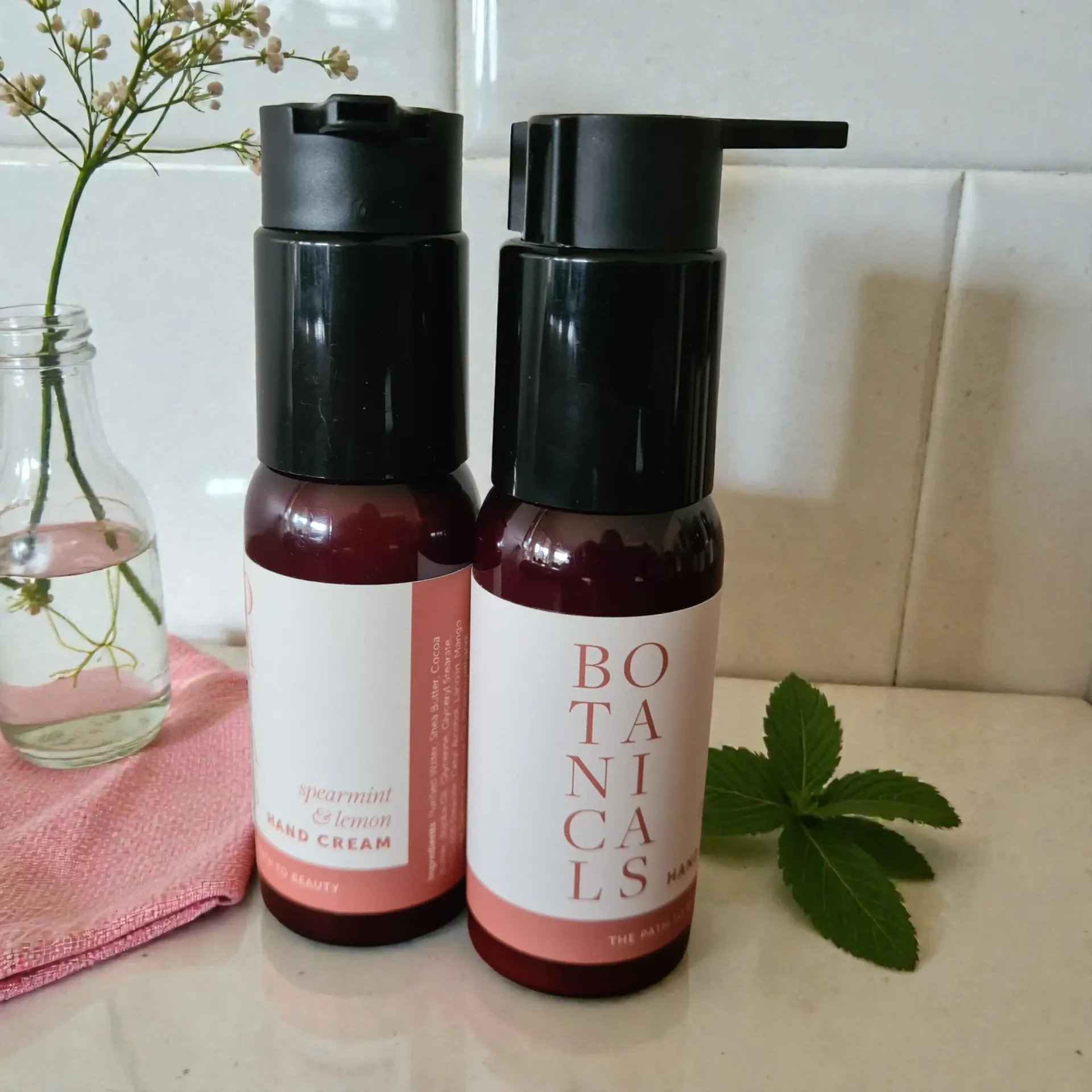 Two amber pump bottles of hand cream with black tops on a tiled counter beside a small vase and green leaves.