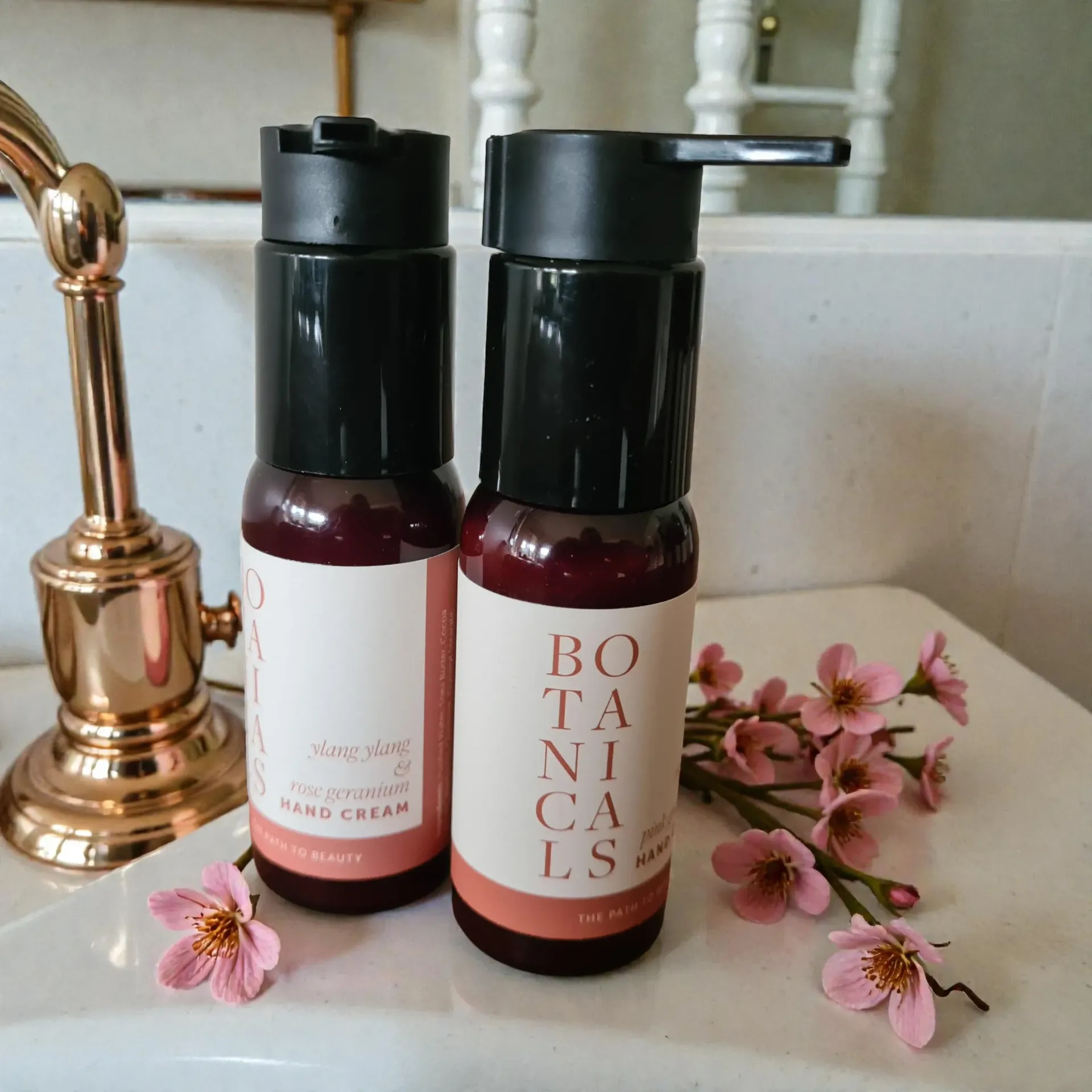 Two pump bottles of hand cream with pink flowers on a white vanity beside a gold faucet