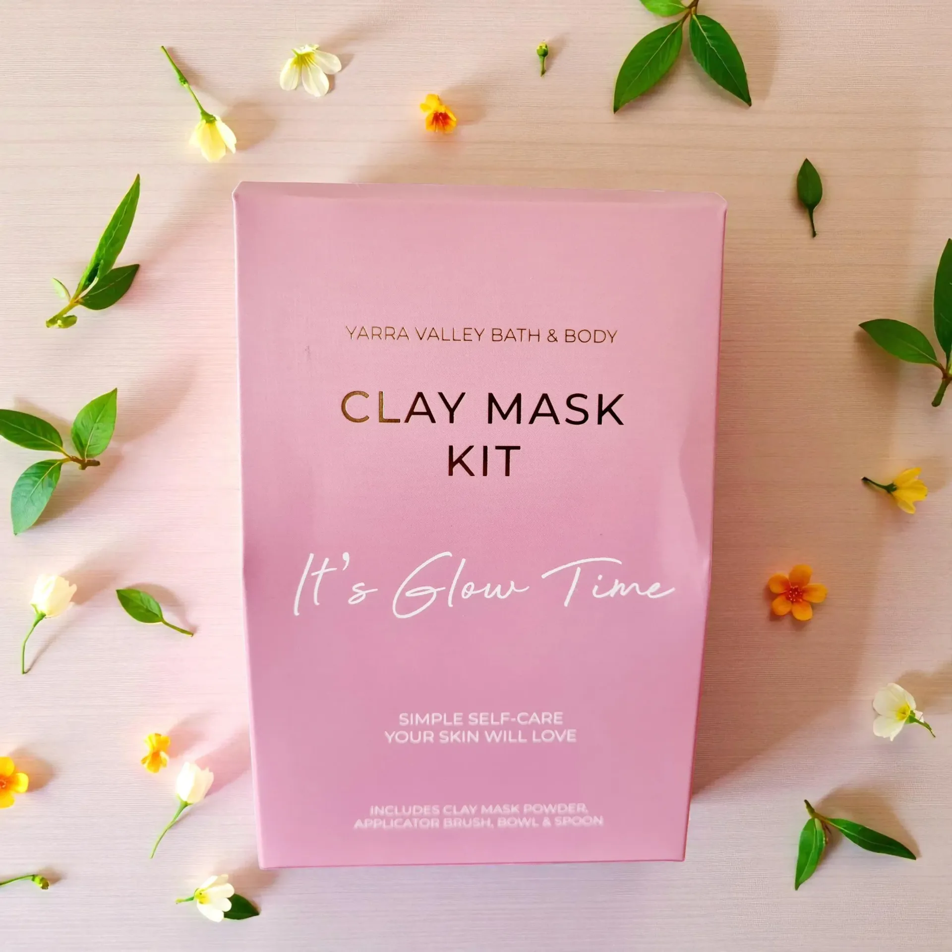 Pink clay mask kit package surrounded by small flowers and green leaves on a beige background