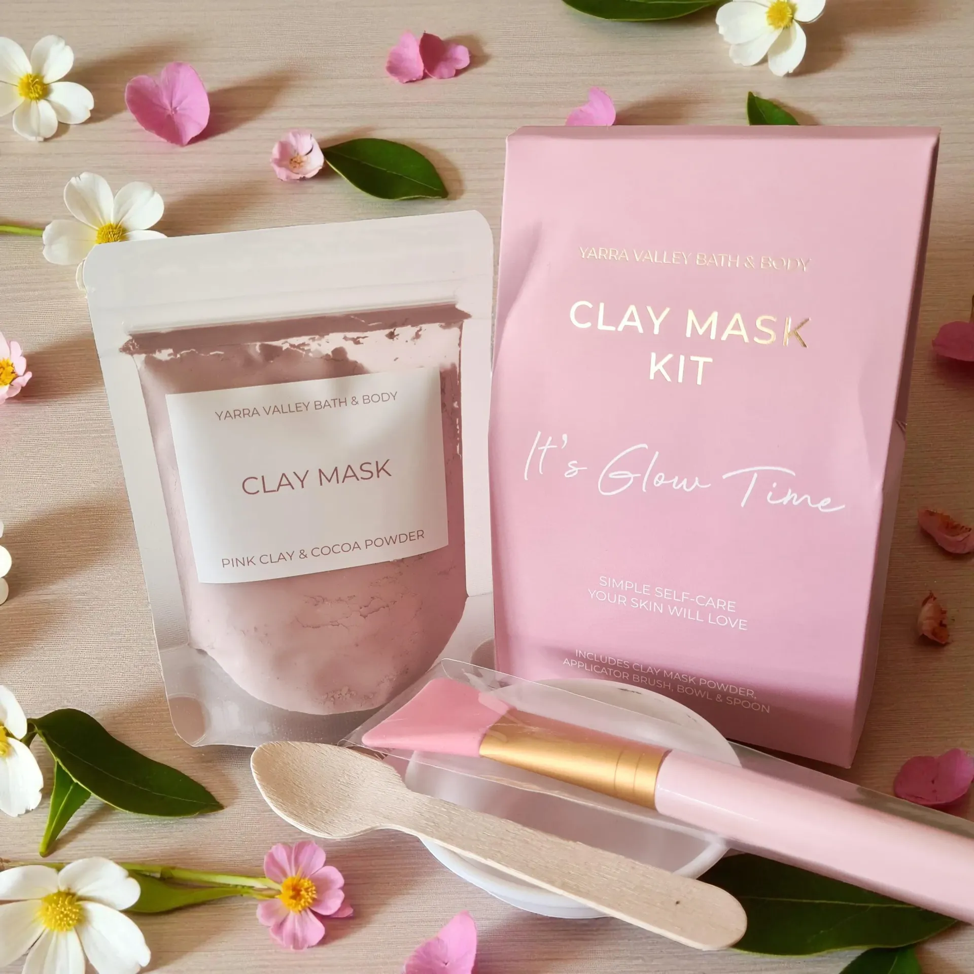 Pink clay mask kit package surrounded by small flowers and green leaves on a beige background