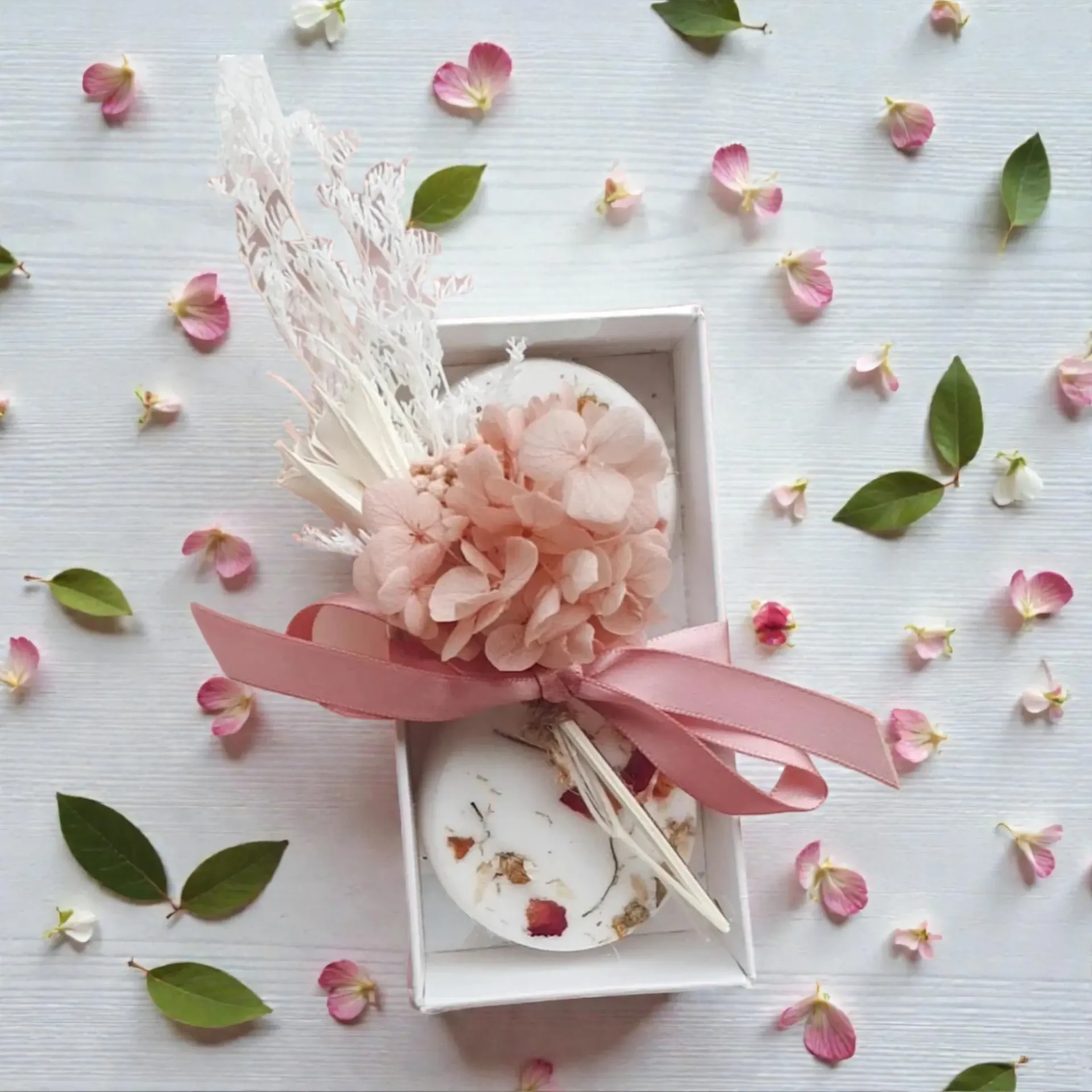 Gift box of shower steamers with pink flowers and ribbon, surrounded by scattered petals and green leaves on a white surface