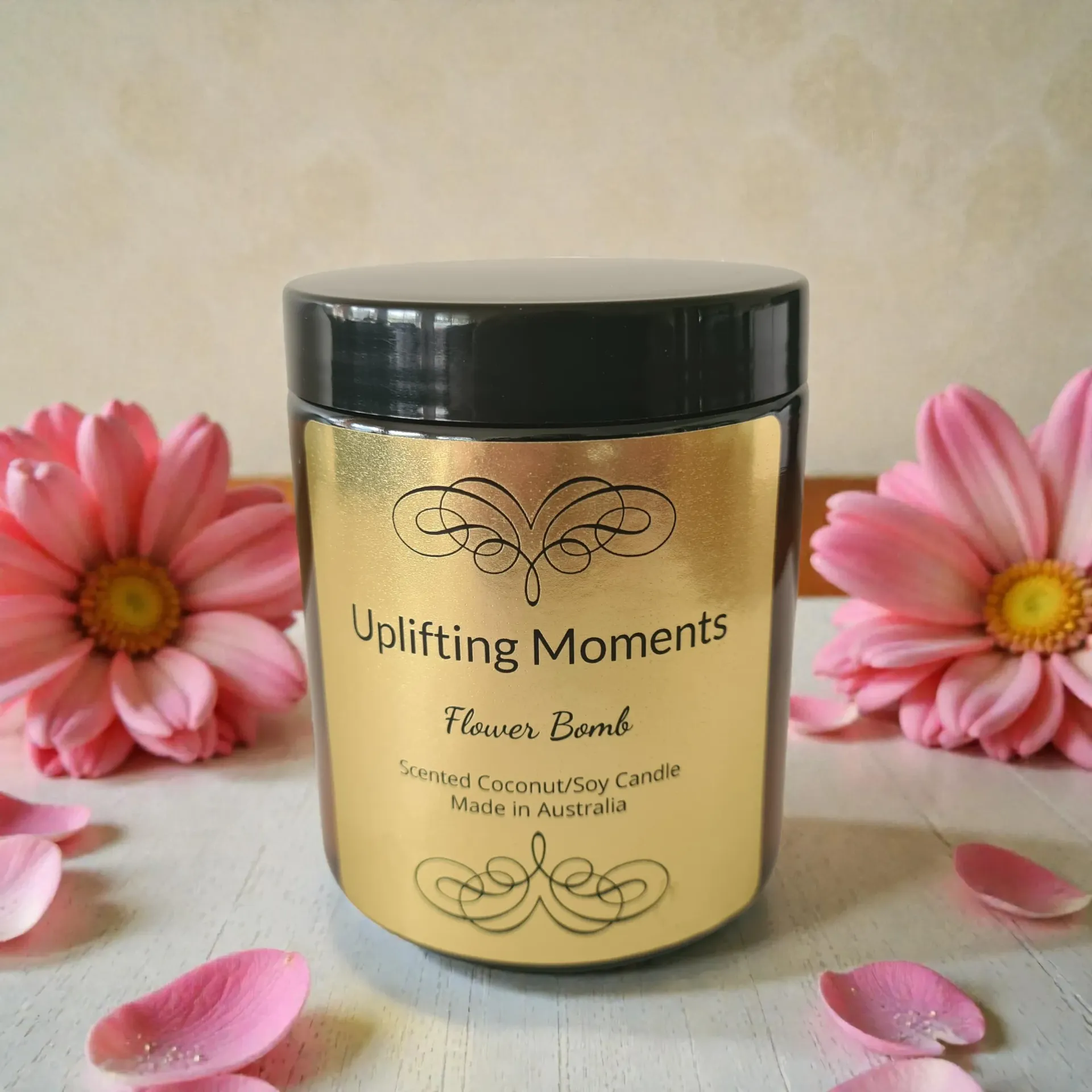 Black jar candle labeled “Uplifting Moments,” surrounded by pink flowers and petals on a soft background.
