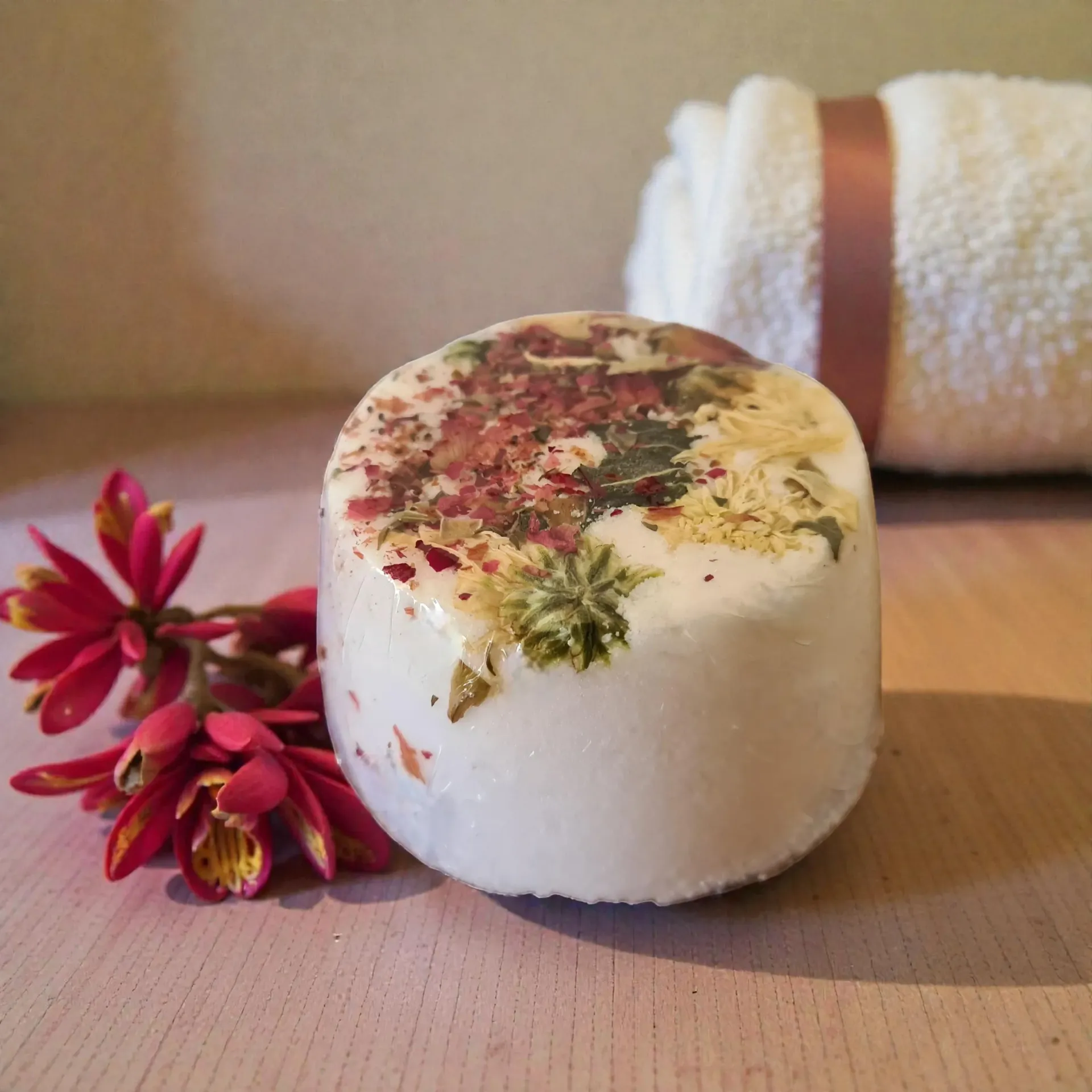 White floral bath bomb on a table with pink flowers and a rolled towel in the background