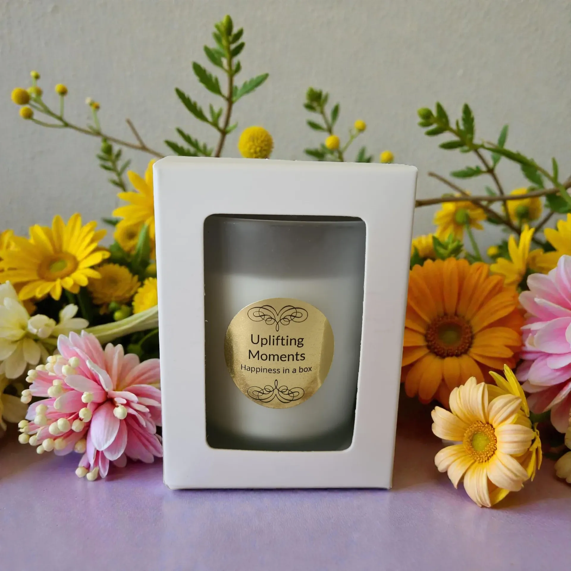 Frosted candle labeled “Uplifting Moments” surrounded by pink, yellow, and red flowers on a wooden surface