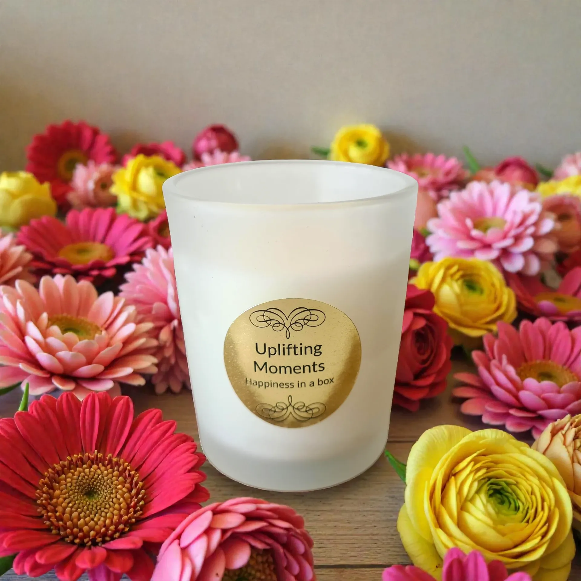 Frosted candle labeled “Uplifting Moments” surrounded by pink, yellow, and red flowers on a wooden surface