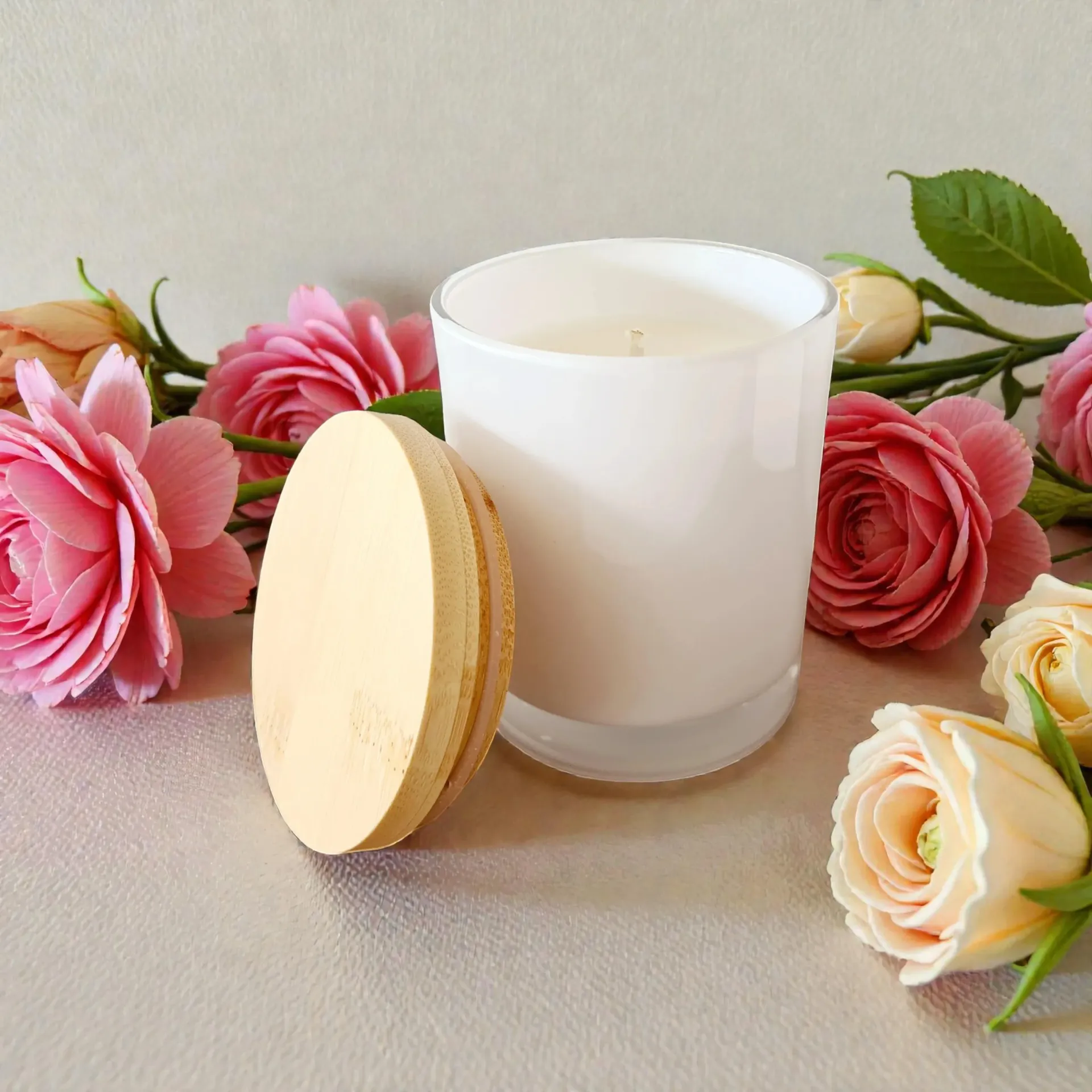 White candle with a round wooden lid, surrounded by pink and cream flowers on a soft background