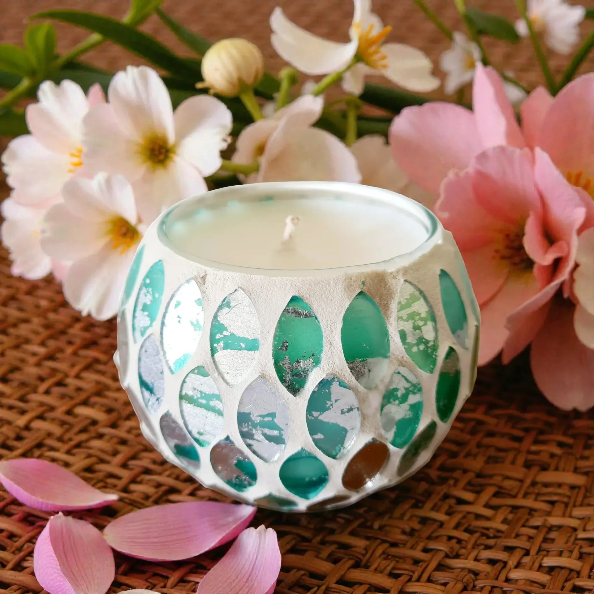 Flower Bomb type candle in a mosaic glass holder surrounded by pink and white flowers on a woven mat