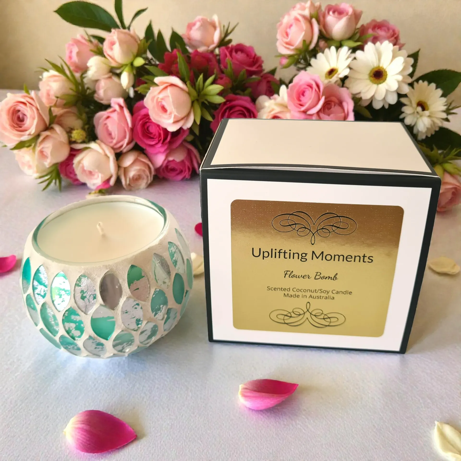 White candle in a mosaic glass holder on woven mat with pink and white flowers nearby