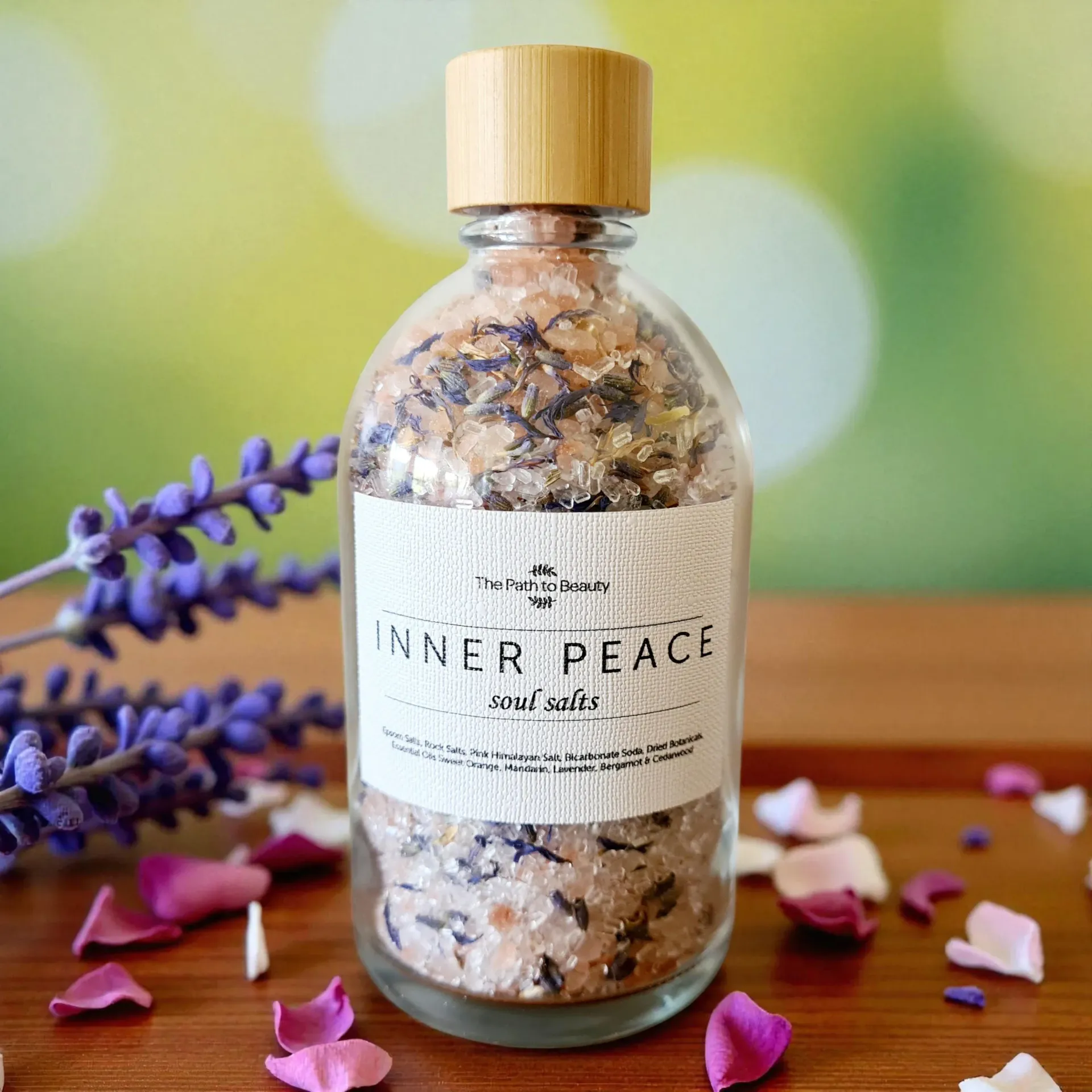 Glass bottle labeled “Inner Peace” with a cork, filled with bath salts on a wooden surface with lavender and petals.