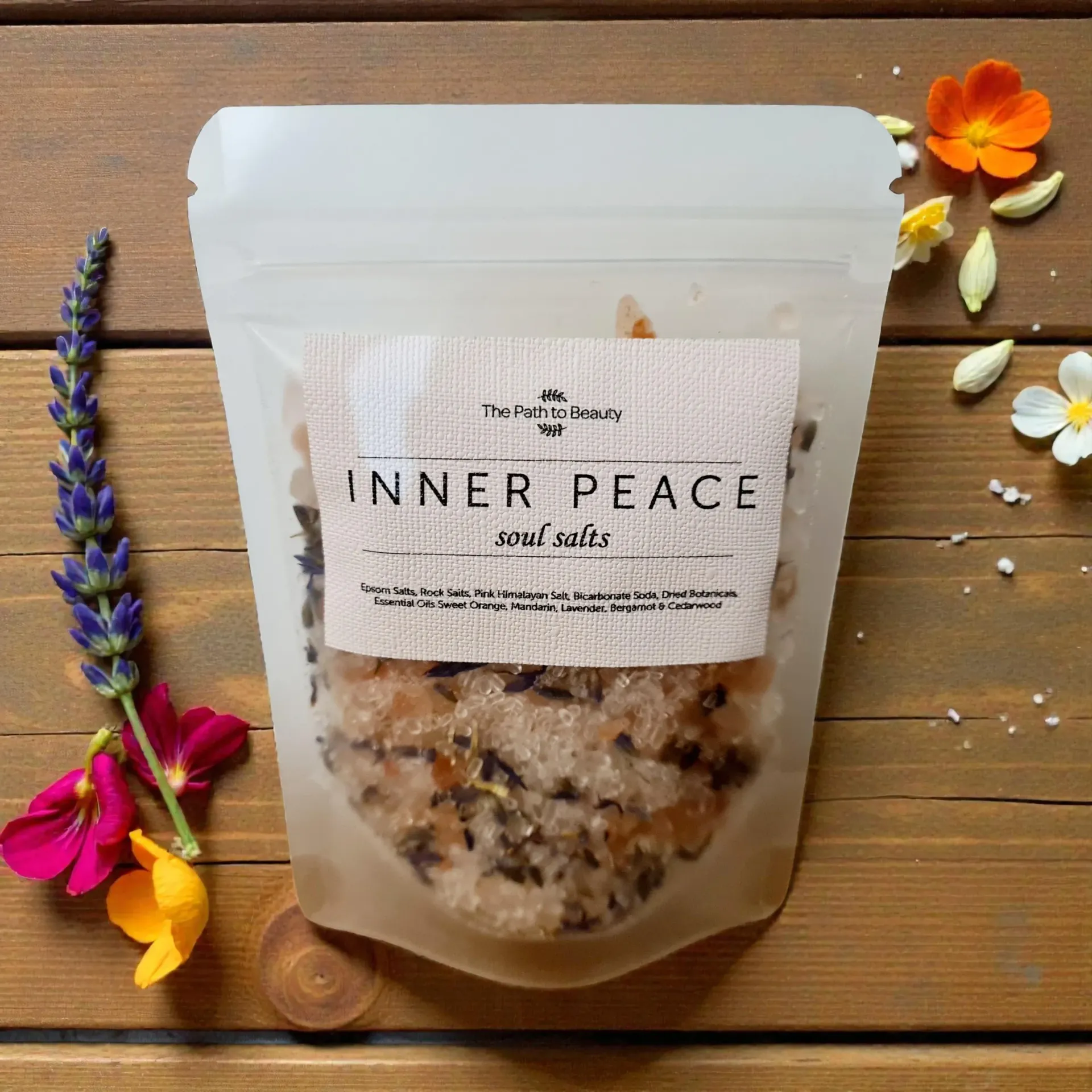 White pouch labeled “Inner Peace” bath salts on wooden table with lavender, flower petals, and orange blossom accents